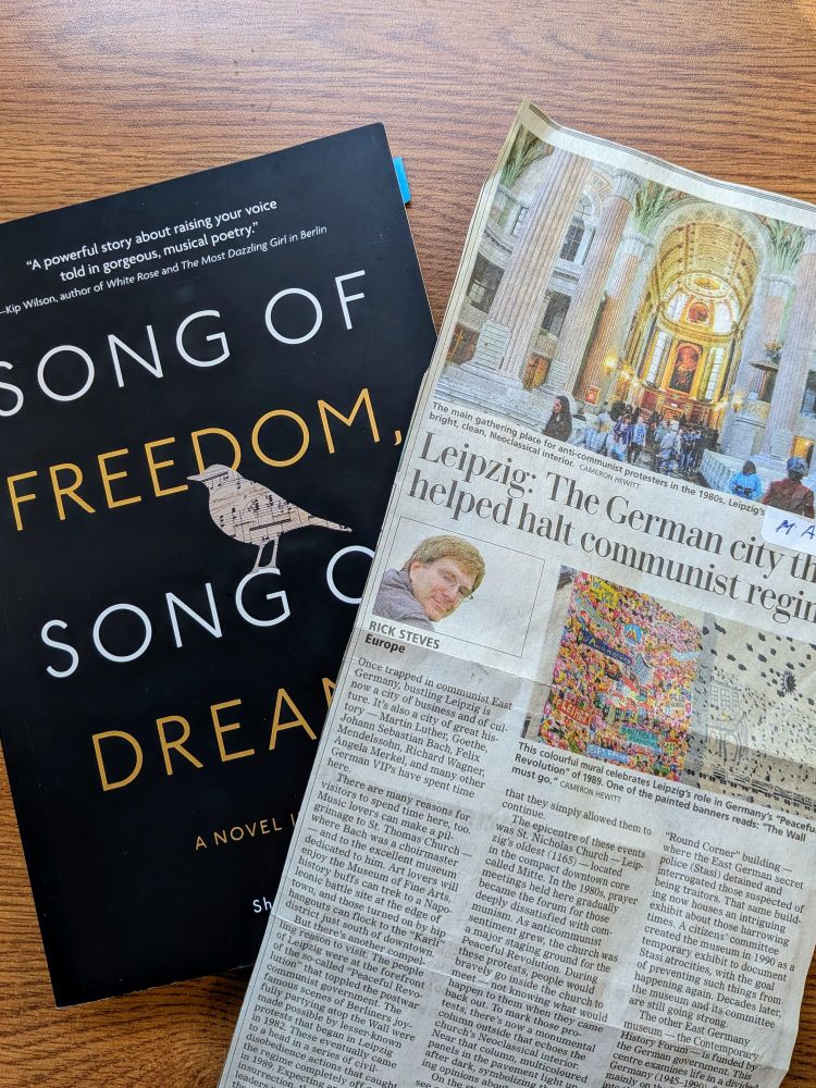 Paperback book with a black cover and white and gold title, "Song of Freedom, Song of Dreams" beside a newspaper clipping of an article by Rick Steves about the German city of Leipzig, with a photo of Leipzig's St Nicholas church (Nikolaikirche).