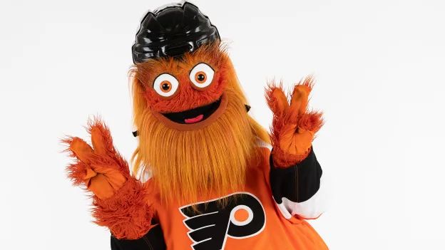 Gritty, giving the (American version) of the v-for-victory sign with both hands. (Large orange hairy mascot of indeterminate gender and species, mascot of the Philadelphia Flyers. They are wearing a Flyers jersey and a black helmet)