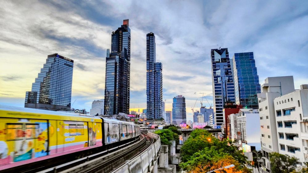 The sky train rolls past above a Bangkok street. Skyscrapers and a big, cloudy sky are in the background 