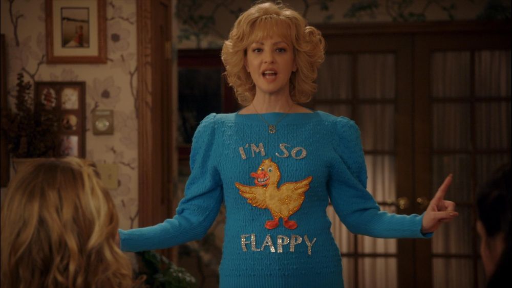 Pic shows the character Beverly Goldberg from the TV show the Goldbergs wearing a light blue jumper with a yellow duck on it surrounded by the words "I'm so flappy" in the living speaking with arms outstretched, index finger on left hand pointing up. A blonde head in left foreground obscures her right hand.