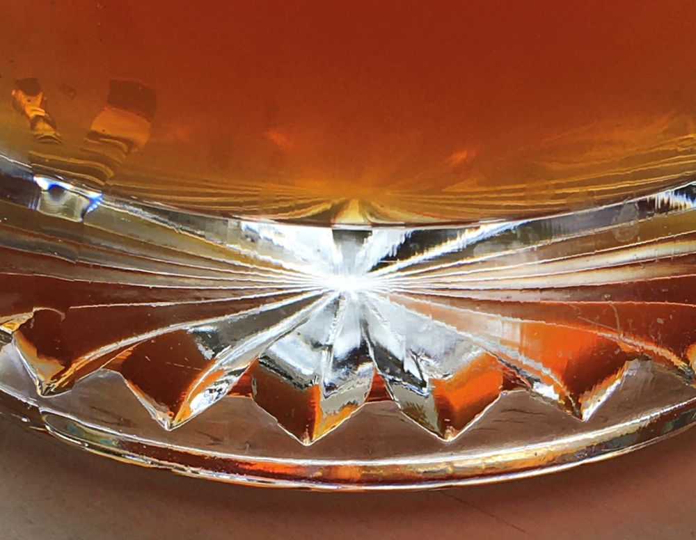 Closeup photo of the bottom of a glass full of golden beer. The glass has an etched design, giving an interesting reflection of the orange liquid emanating from a white/silver center.