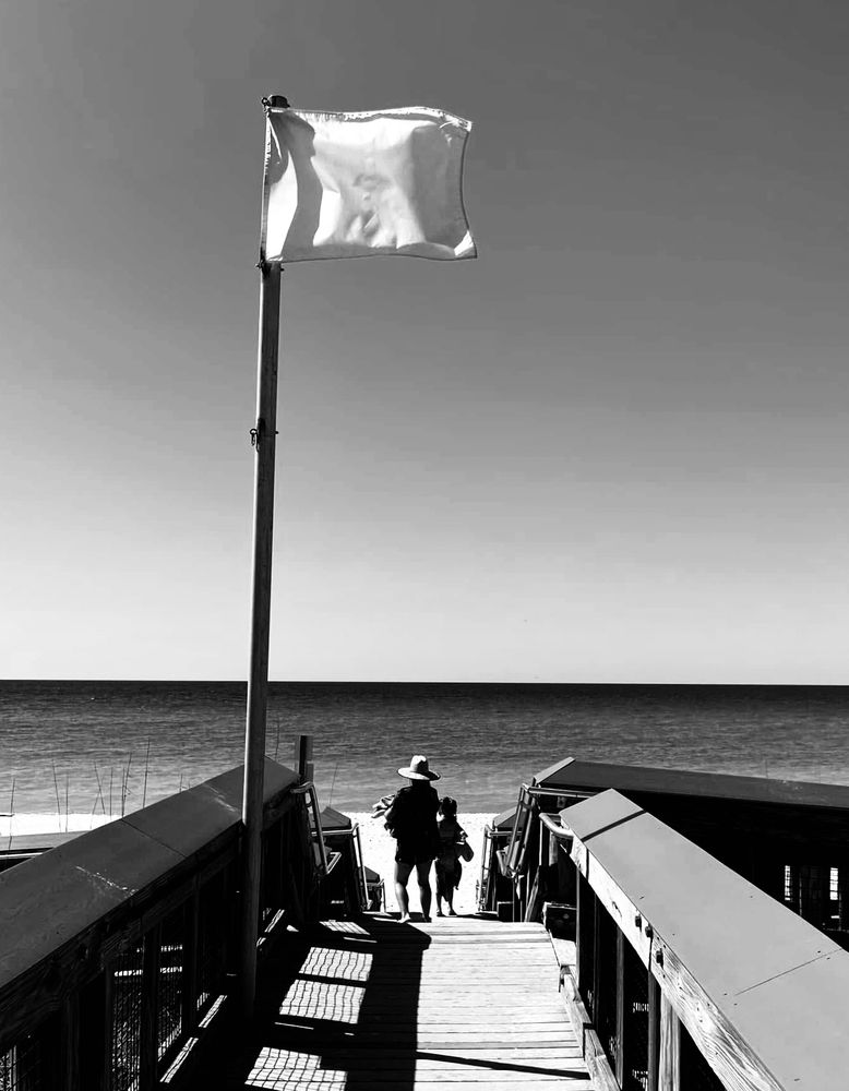 Black and white photo looking down a wooden bridge to the sand and sea. A tall pole with a flag atop is on the left side of the bridge. Two people are at the end of bridge.