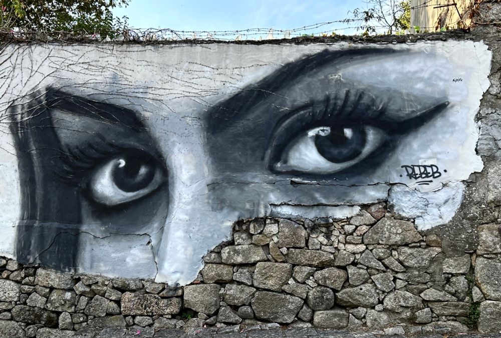 Photo of a black and light gray painting on a beige stone wall, of the top of a ladies face. Large black eyes, with eyeliner above and below eyes, and nice arched black eyebrows. 