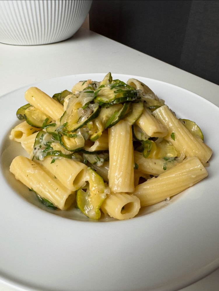 A shallow bowl with long, ribbed pasta cylinders tossed with cooked zucchini and chopped parsley.