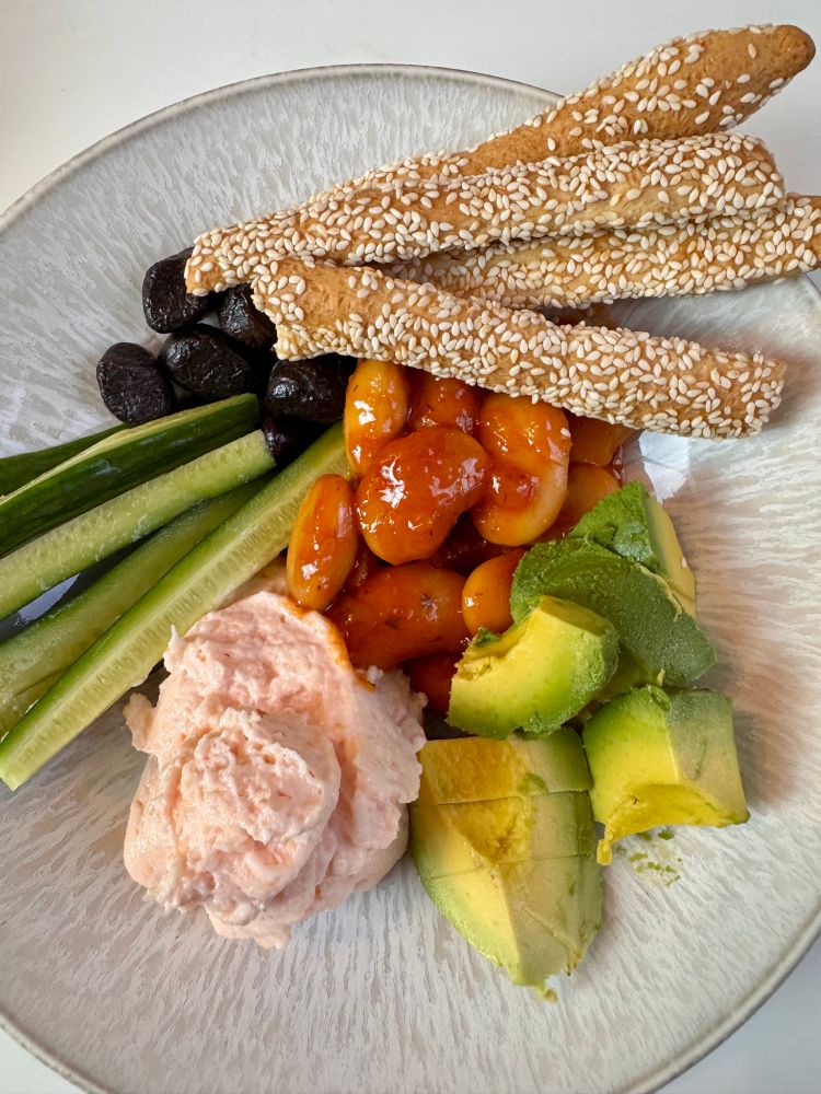 A summer cold plate with pinkish cod roe dip, chunks of light green avocado, Persian cucumber spears, ink-black olives, tomato red marinated gigante beans, and crisp and golden sesame sticks.