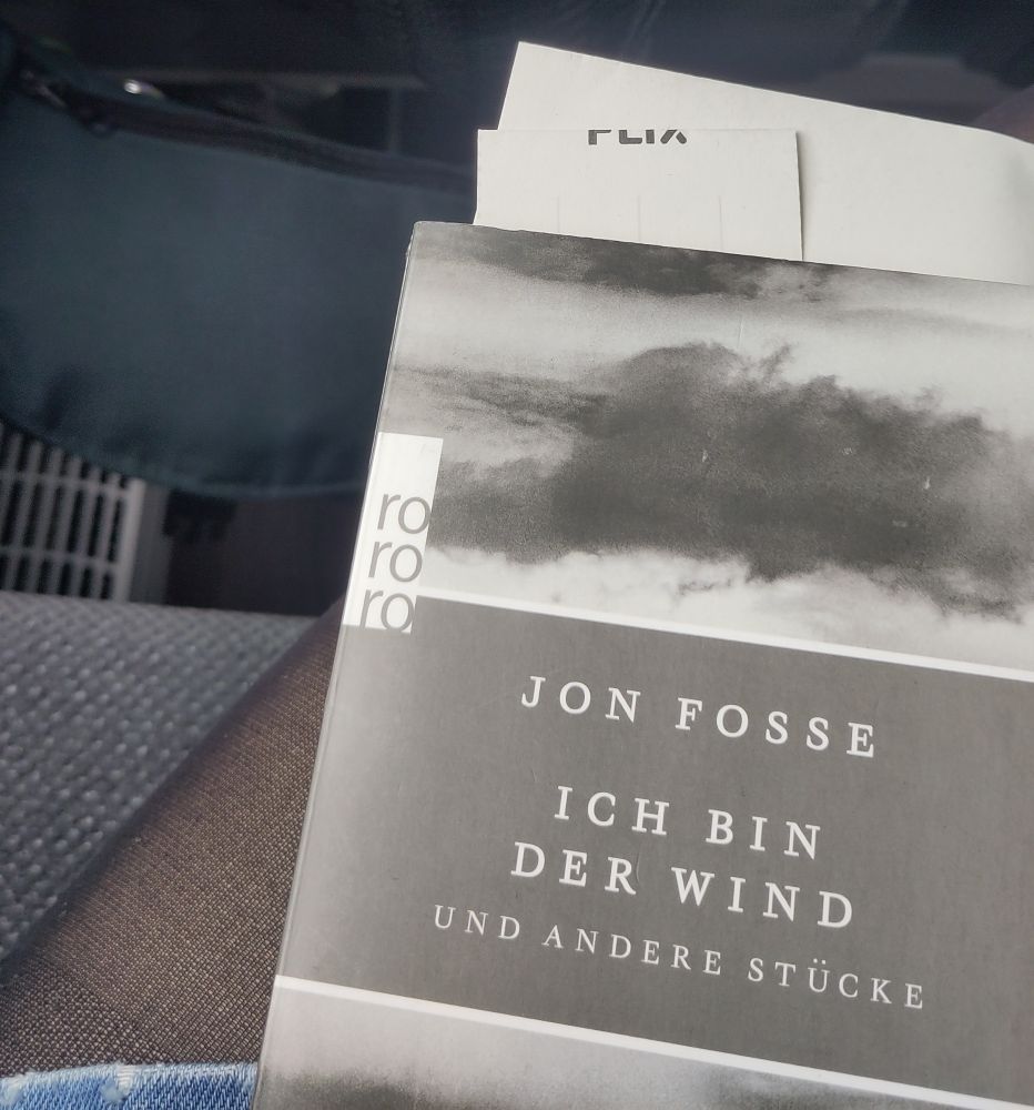 Im Flixbus, man sieht das Buch Ich bin der Wind und andere Stücke von Jon Fosse, das Buchcover ist grau mit grauen Wolken