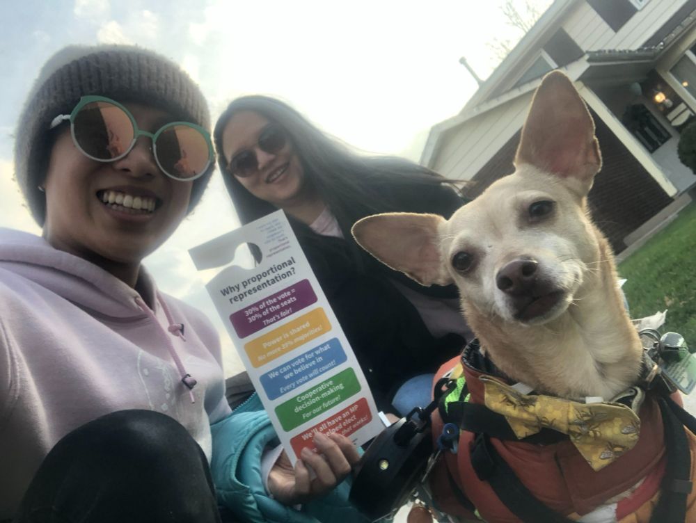 Two smiling volunteers pose for a selfie while sitting on a lawn, holding a Fair Vote Canada door hanger that lists reasons to support proportional representation. A small dog in a yellow bow tie and orange vest is also facing the camera.