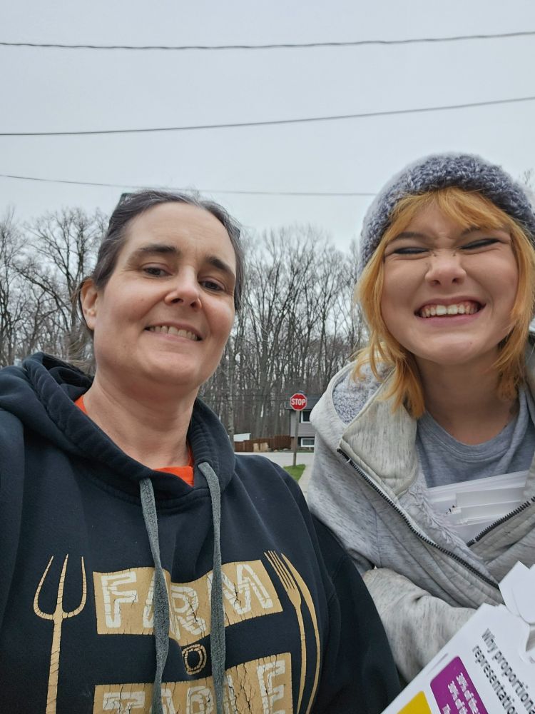 Two volunteers smile at the camera while out delivering door hangers. One wears a “Farm to Table” hoodie, and the other, holding a stack of Fair Vote Canada flyers, wears a beanie and a grey hoodie. Leafless trees and a stop sign are visible in the background.