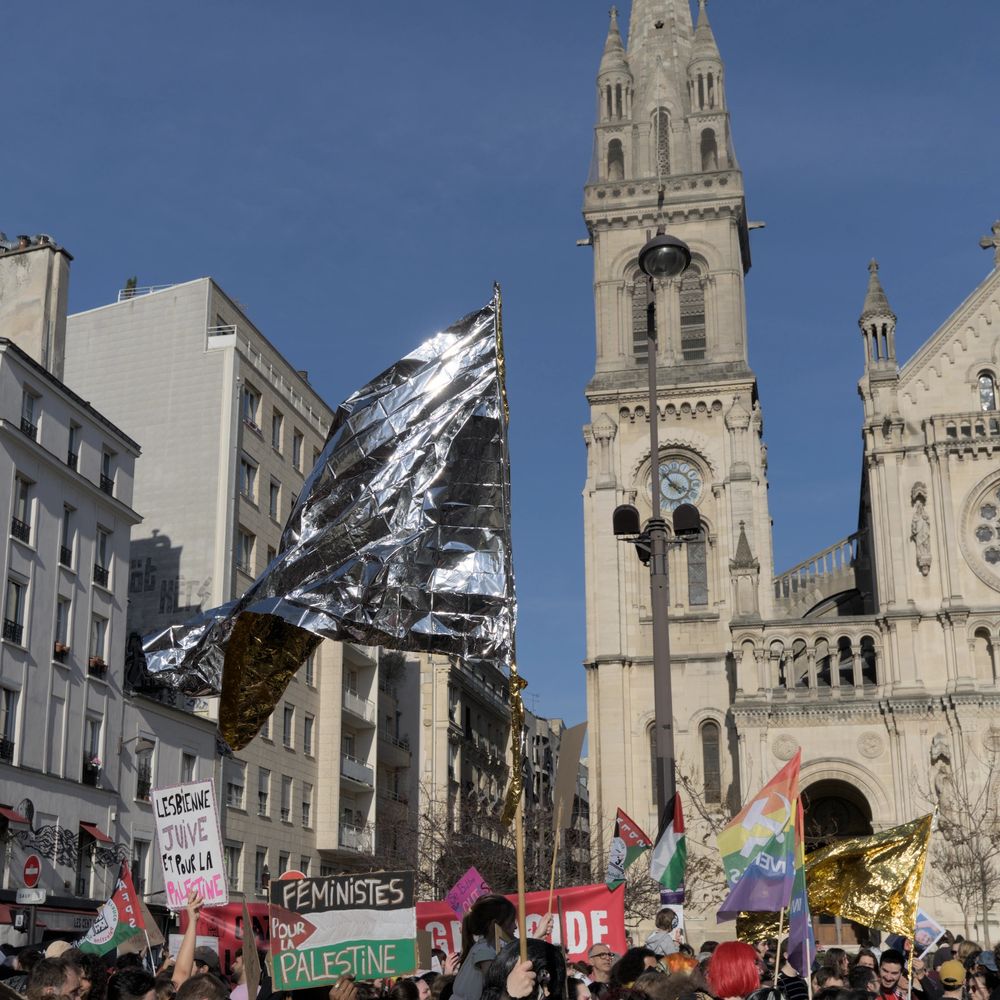 Flags and signs of the march above protesters' heads in front of the Saint-Ambroise church in Paris. The center of the image is a huge silver flag made of tin-foil, reflecting the sunlight. Other signs include "Feminists for Palestine" on a Palestinian flag, "Jewish Lesbian and for Palestine" (both translated from French), rainbow flags with a hammer and sickle, a golden metallic foil flag.