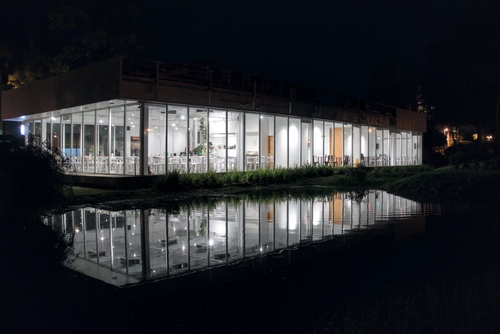 Photograph of a coffee shop building in the night, by a pond. The coffee shop interior is the only source of light. It is a single-storey, modern building lighted with bright, crisp white light. The sides of the building facing the photographer are all glass and the deserted building interior can be fully seen.
Its facade and its white light are reflected in the pitch-black pond separating it from the photographer.