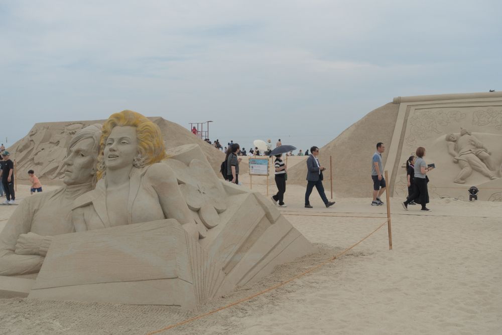 Photograph of a sand sculpture exhibition on a beach. The preeminent sculpture in view represents the busts of Andy Warhol and Marilyn Monroe. The sand for Marilyn Monroe's hair is colored yellow.