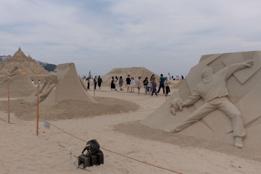 Photograph of a sand sculpture exhibition on a large beach. The nearest sculpture to the camera represents a masked man in the act of throwing a Molotov cocktail. Further in the background, another sculpture represents the character from Edvard Munch's Scream. Other sand sculptures can be seen farther away.