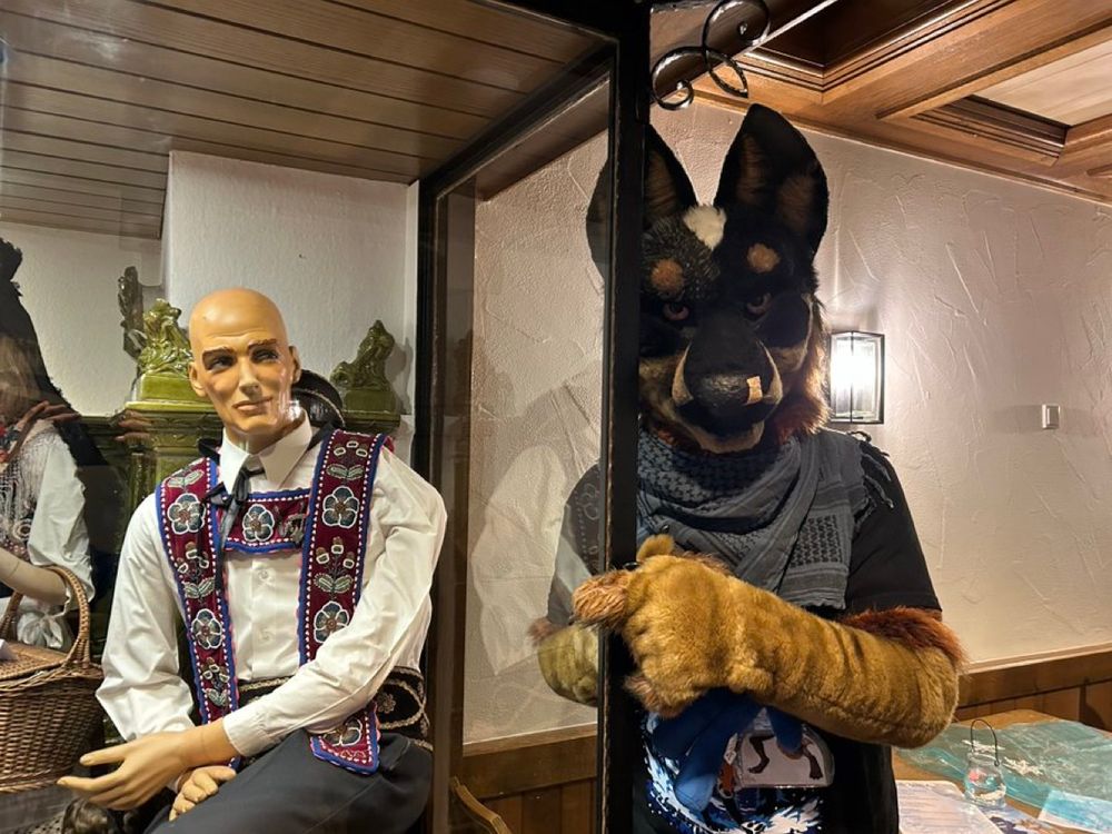 Photo of a Blue Heeler fursuiter next to a mannequin looking remarkably similar to Agent 47 from the Hitman game series.