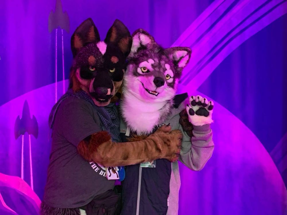 Wolf and Blue Heeler fursuiters in front of a space-themed background, the Blue Heeler fursuiter hugging the wolf fursuiter.