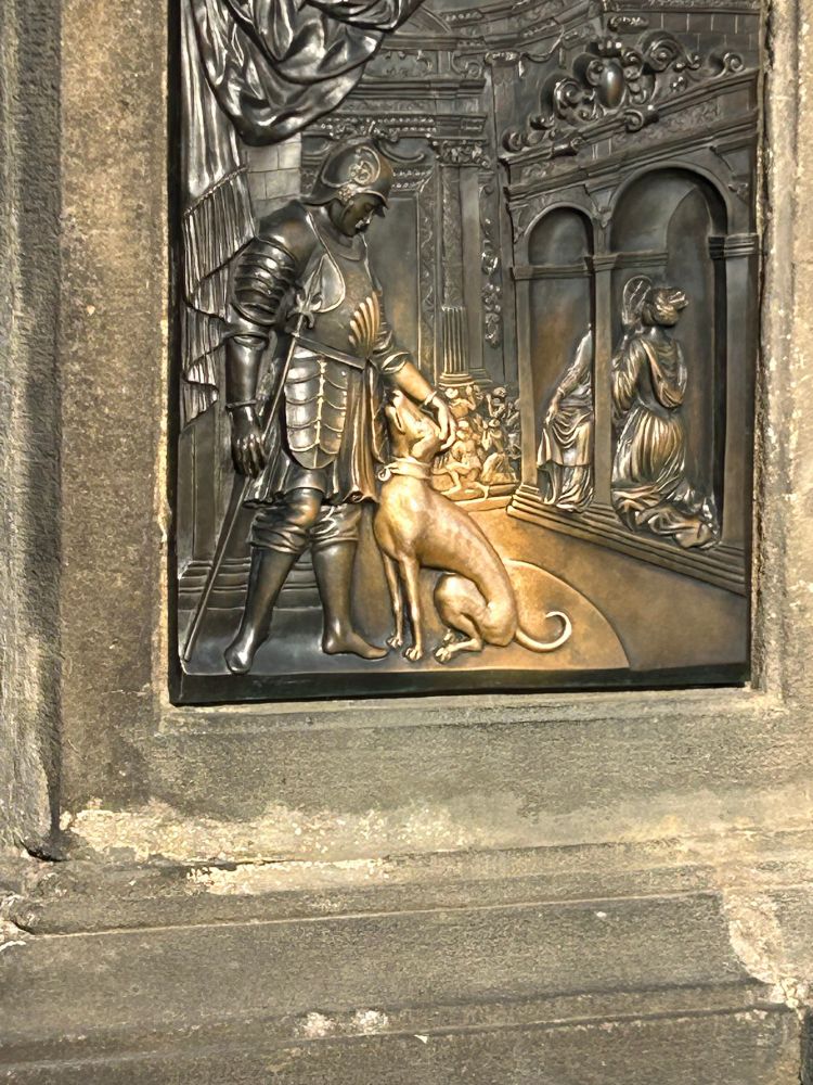 Photo of a relief on the socket of a statue at Charles Bridge, Prague, depicting a dog which is shiny and clean in contrast to the rest of the relief due to visitors petting it.