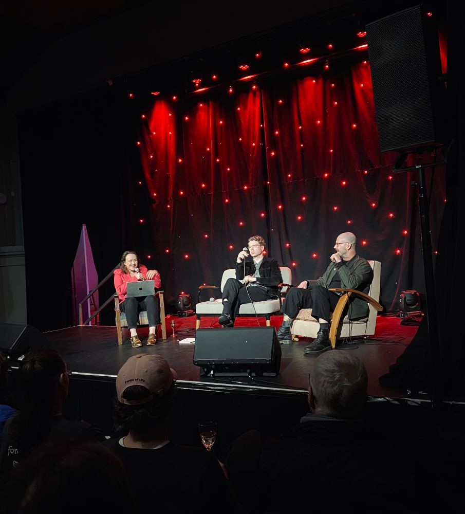 A photograph of three people on stage sitting in armchairs holding microphones. They are discussing a book for its launch. The backdrop is filled with little red twinkling lights. 