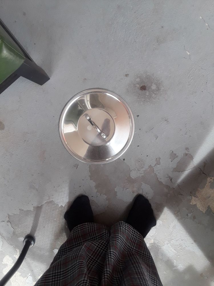 A stainless steel lid for a soup pot on a concrete floor. It simply appeared there. I have no idea how it arrived, when, or why it's on a covered balcony 8 stories up. Also pictured are my confused feet and the legs of random chairs