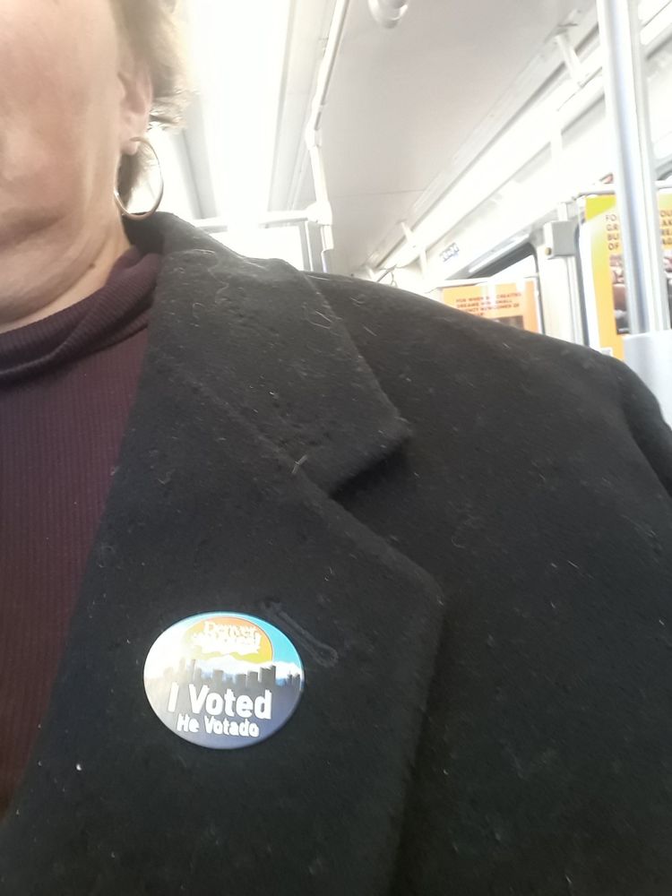 A round sticker with a light blue sky, a mountainscape, and a skyline in darker blues, with a golden sun circle saying Denver votes.
Below is printed I voted. The sticker is on a black coat covered in lint, over a purple turtleneck. Above is a bit of my neck, and my double chin is more evident than I'd like it to be, but I also like candy