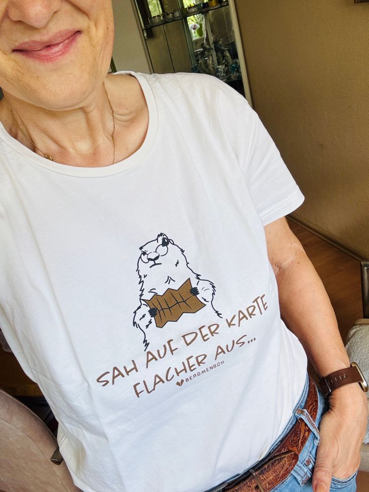 Eine Frau mit einem beigen Shirt. Darauf liest ein Murmeltier eine Landkarte. Darunter steht „Sah auf der Karte flacher aus“ und die Marke „Bergmensch“