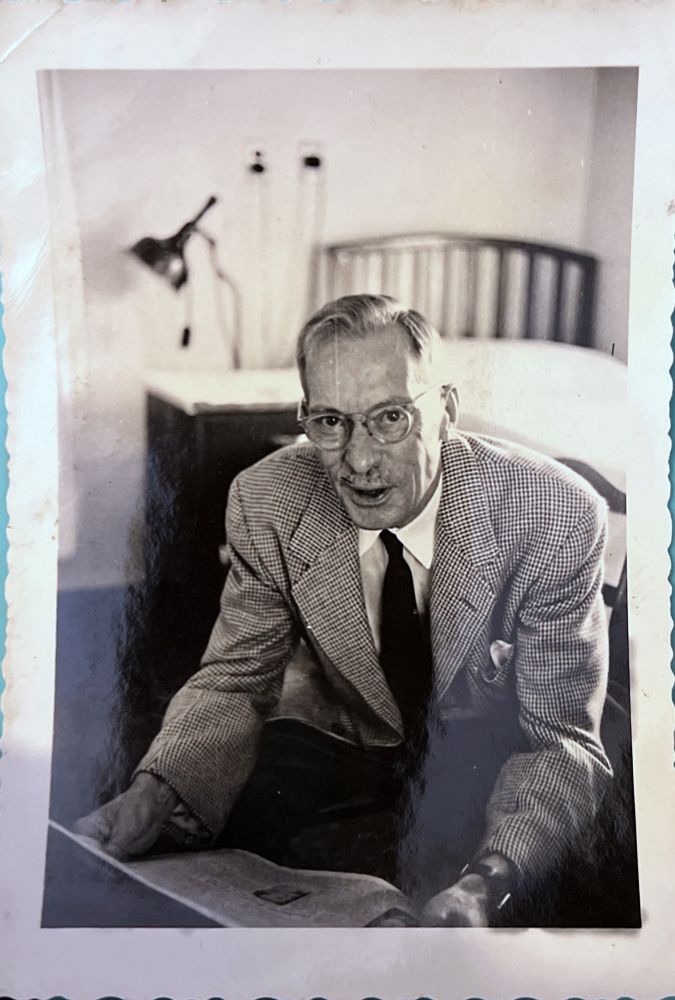 An old black and white photo showing an older man sitting in a chair. He is looking at the camera. He has round glasses and a slight mustache. His mouth is open in a sort of surprised smile. He is wearing what looks like a tweed or checked suit jacket, white dress shirt, and black tie. He is holding a newspaper and the background has a slightly blurred bed and nightstand.