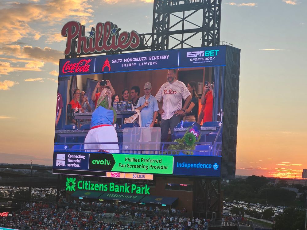 A photo of the Phillies board, which is showing the Phanatic rolling on the ground after being caught with another Phanatic in a blond wig. She is hiding her face from the camera. 