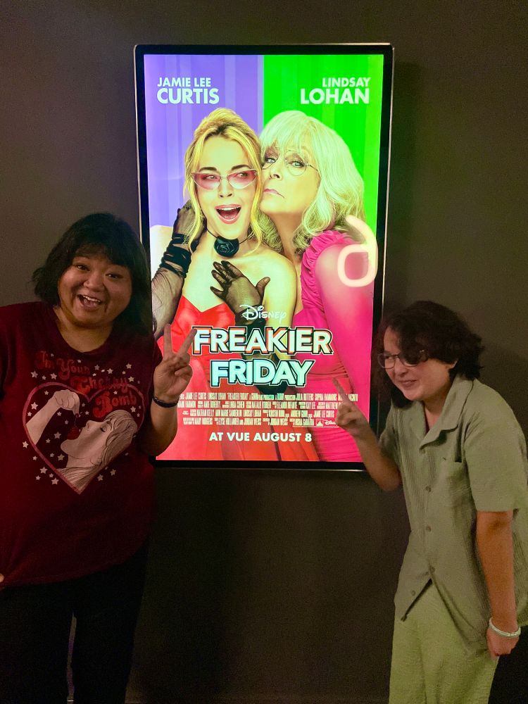 Me and my 12 year old daughter either side of the ‘Freakier Friday’ poster of Lindsay Lohan and Jamie Lee Curtis. She’s looking sideways at me and grinning while waggling a finger and I’m smiling right at the camera and doing the peace sign. She’s dressed in a nice teal outfit with a buttoned shirt and I’m wearing a T-shirt that says ‘I’m your Cherry Bomb’ it’s an illustration of a woman eating a cherry, surrounded by a heart and stars.