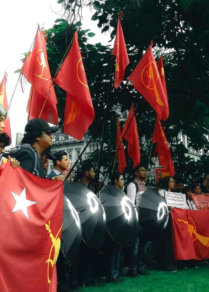 A group of people standing side on, holding red NLD flags (Myanmar’s National League for Democracy - the rightful but exiled ruling party) and four black umbrellas each with number 8 written in Burmese (၈၈၈၈)