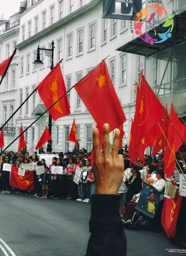 More protestors/marchers holding placards and red NLD flags. In the foreground you can see the hand of someone doing the three finger salute, the symbol of resistance.