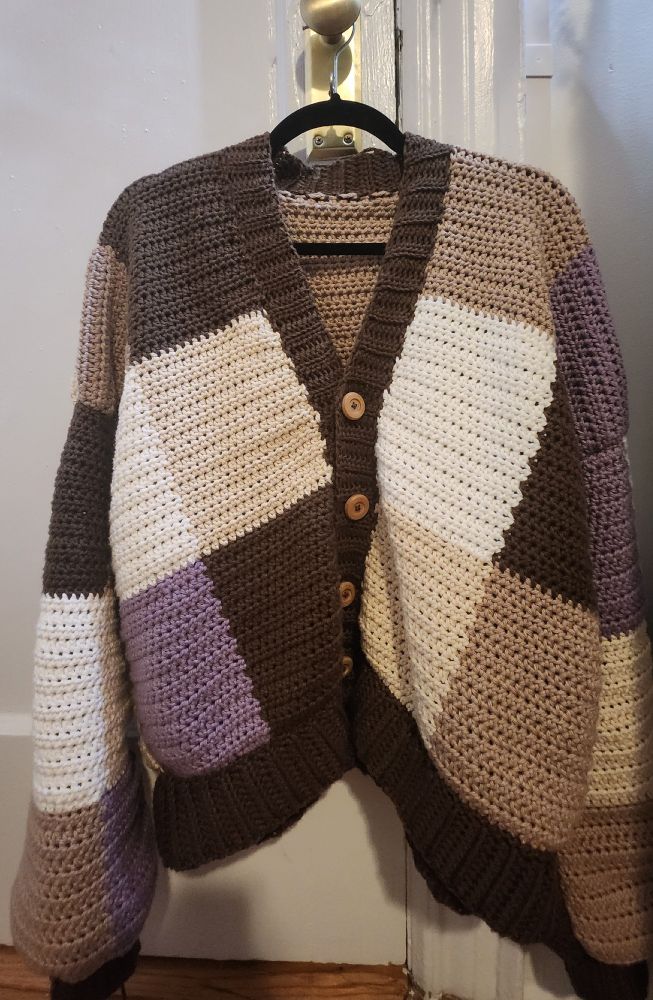 A crocheted cardigan on a hanger with alternating color squares of purple, white, brown, beige, and tan combined with brown ribbing and light brown buttons