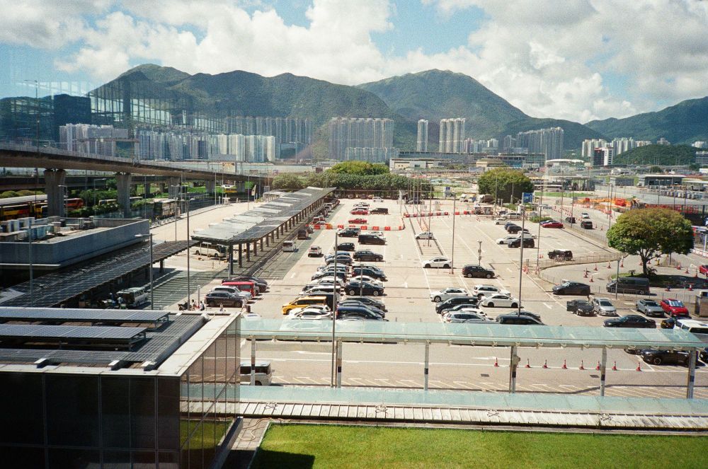 A scene from out the window at Hong Kong Airport. There are cars parked and beyond that, buildings and mountaintops. There are some reflections coming from inside the terminal