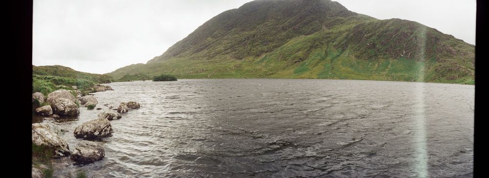 A large lough with a mountain in the background. The sky is overcast and seems pure white from this angle. There are old boulders in the foreground