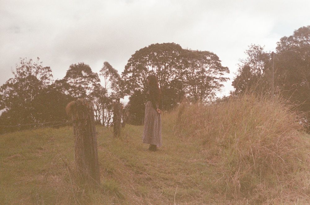 a woman is staring off in the distance in the middle of the frame, she is standing in a field, the image is extremely grainy due to using expired film