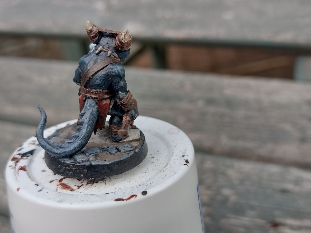 Rear view of a greyish blue pathfinder style kobold, holding a staff and conjuring something in their left hand.