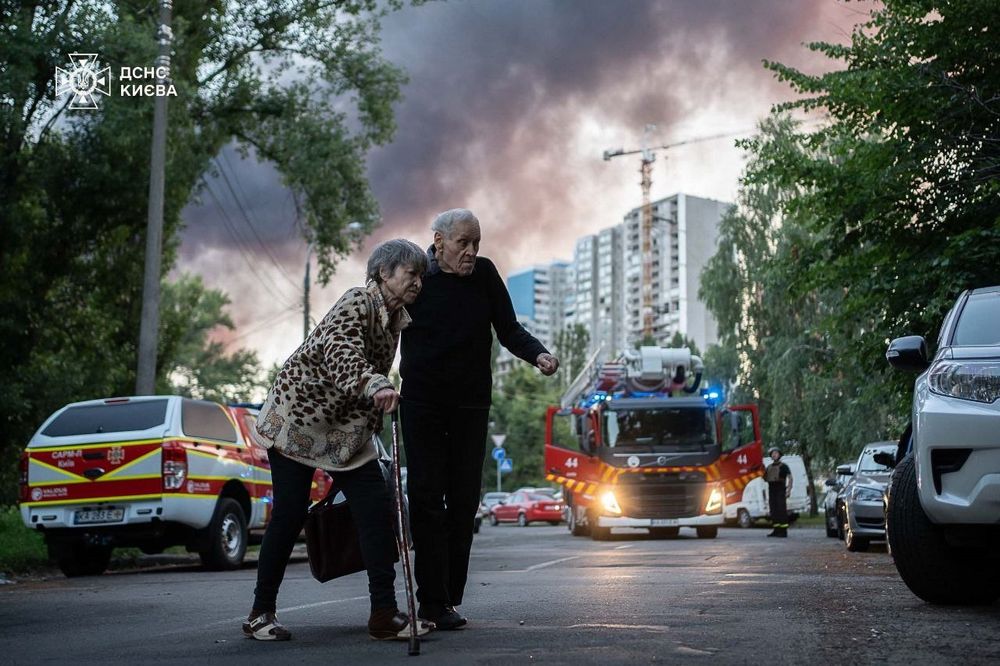A couple crossing the road in Kyiv, with rescuers in the background in a firefighter vehicle, smoke and a residential building behind.