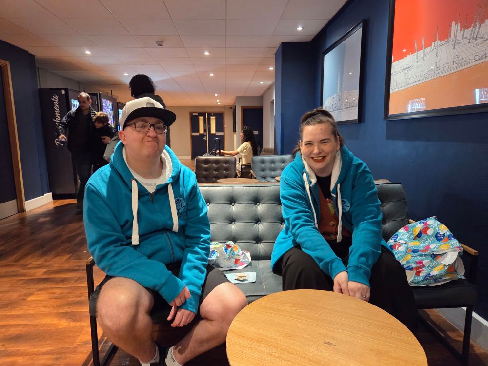 Mat and Harriet in aqua blue teal hoodies sat on a sofa in hotel lobby. They smile like happy children.