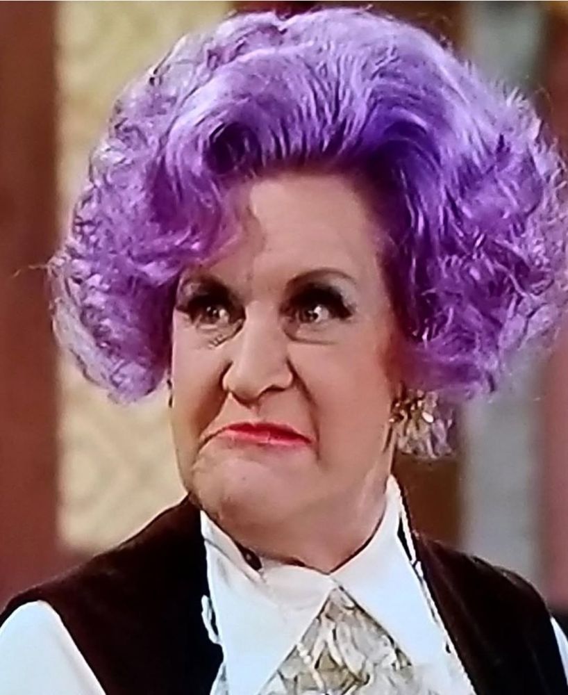 A photo of actress Mollie Sugden in the role of Mrs. Slocombe from the BBC comedy Are You Being Served? She has violet-colored hair and is scowling. 