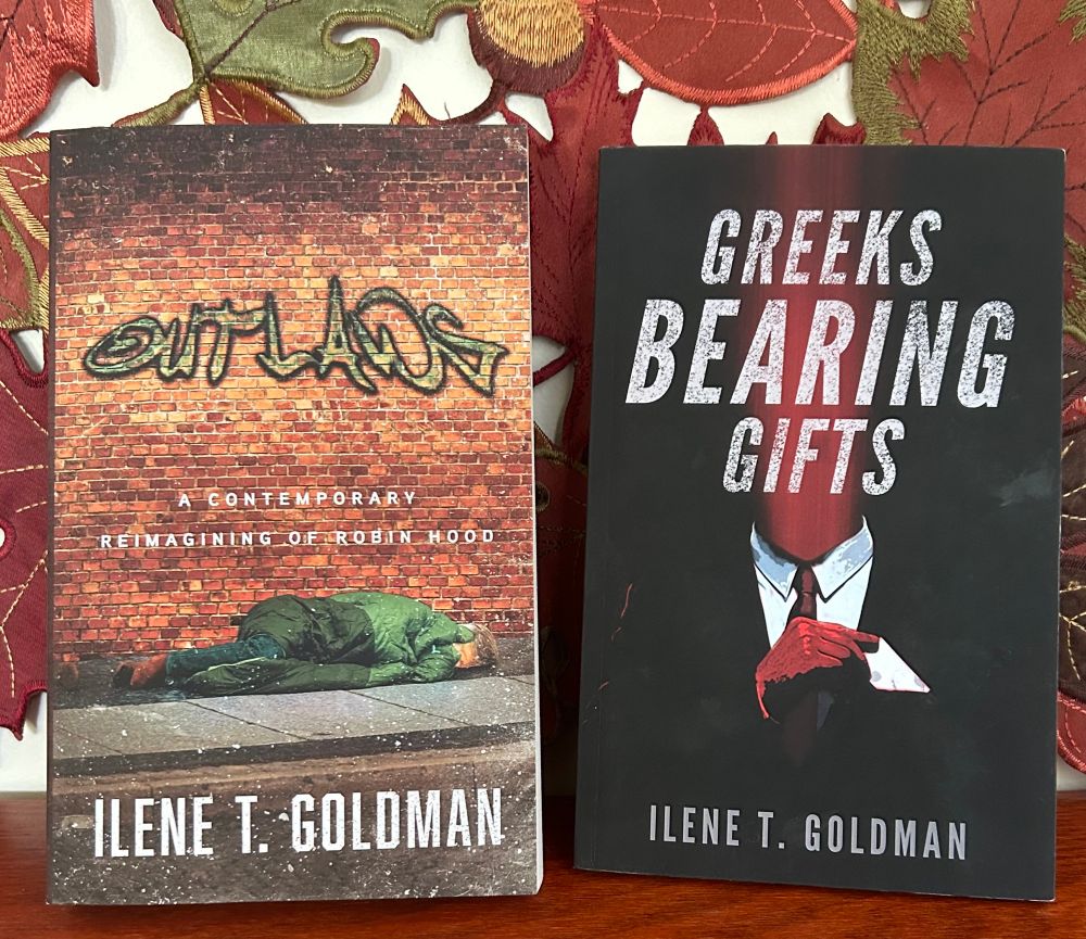 Two paperbacks against an autumn leaves background: Outlaws: A Contemporary Reimagining of Robin Hood and Greeks Bearing Gifts, both by Ilene T. Goldman