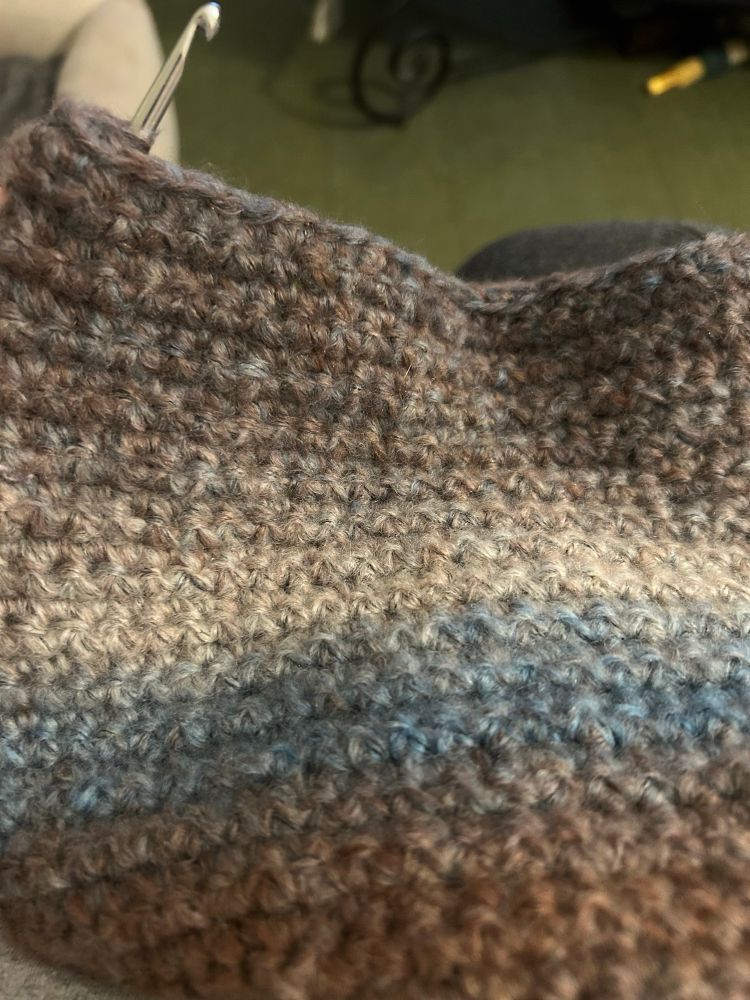 A grey, teal, and brown scarf-in-progress. A crochet hook sticks through the top right corner. 