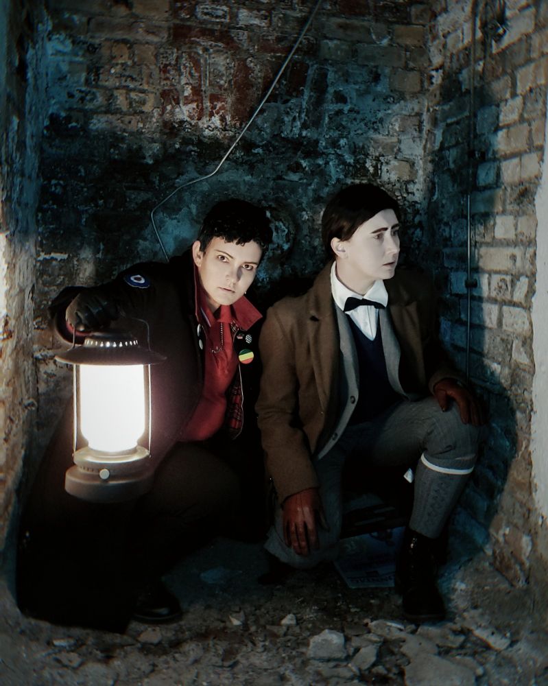 Zwei Cosplayer verkleidet als Edwin Payne und Charles Rowland aus Dead Boy Detectives knien in einer dunklen Nische. Charles hält eine Lampe und beide sehen sich misstrauisch um.