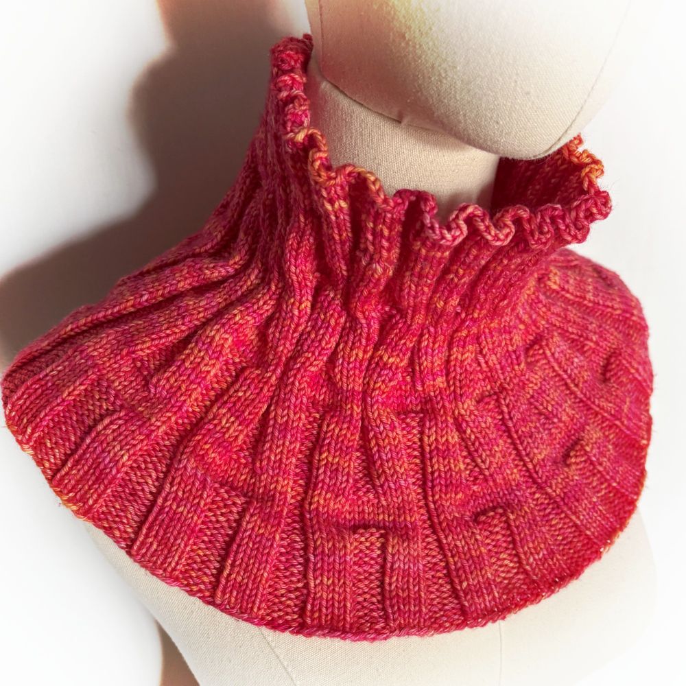 A hand knit cowl on a natural tone linen mannequin with a white background with shadow. The cowl has a textured ribbing pattern of 'missed' ribs that create divots or dips in the fabric and make the ribs appear to lean into each other. The pattern is @LavishCraft Rib and Divot Cowl. Ravelry https://www.ravelry.com/patterns/library/rib-and-divot  or 
PayHip https://payhip.com/b/PQC13
It is worked in a bright pink and orange tonal kettle dyed yarn. Malabrigo Rios in Gemini.
(This is not a shaped item, it has lots of negative ease and flex to fit over the upper chest but still stand up around the neck on it's own)