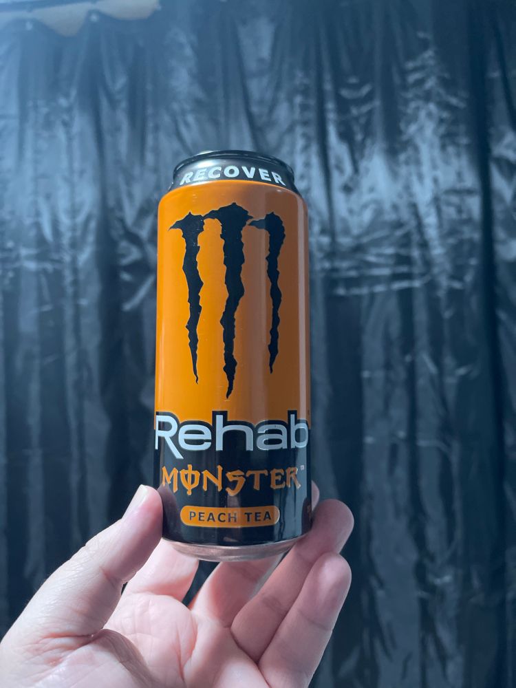 A hand holding up a can of Monster Rehab, Peach Tea” energy drink in front of a black curtain
