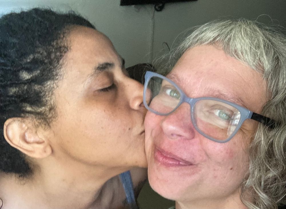 Close up of a brown non-binary person with black dreadlocks kisses a white woman with white hair in blue glasses on the cheek. The white wan smiles at the camera