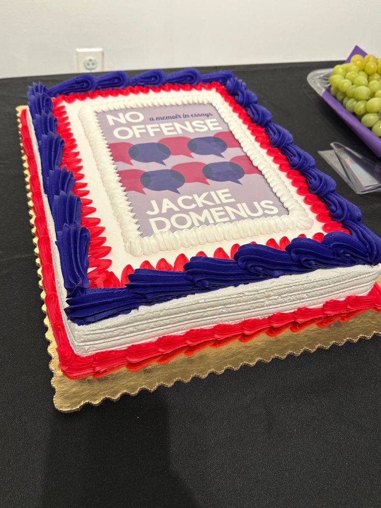 A book cake with the cover of No Offense printed on it surrounded by purple and pink icing.