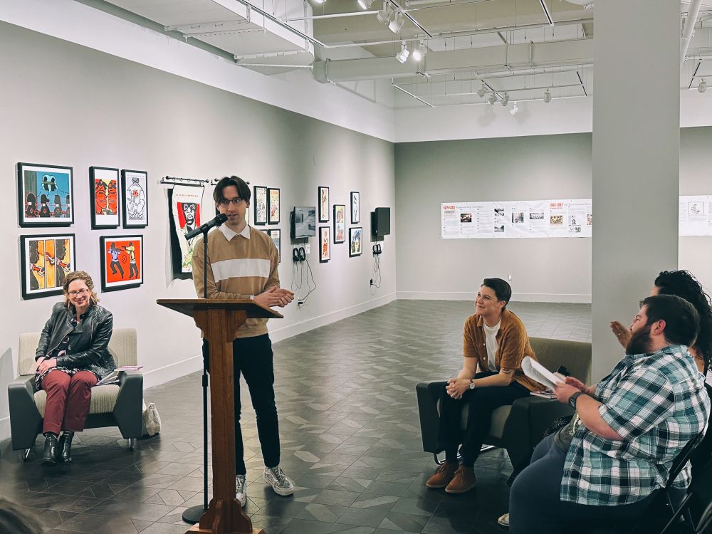 Author Matthew Vesely standing at a podium speaking into a microphone as others watch from chairs behind him.