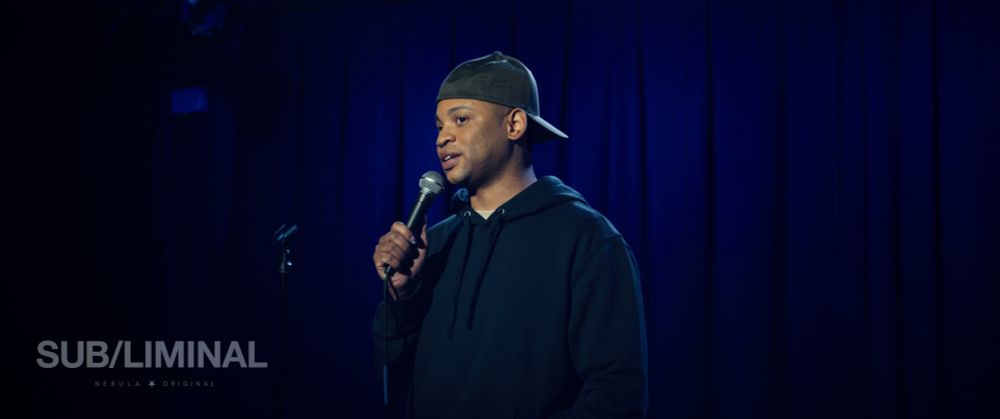 Ryan Jamaal Swain stands in front of a blue curtain holding a microphone and wearing a baseball cap backwards.