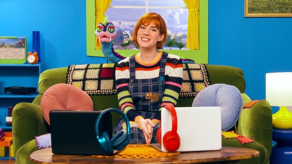 Maggie Mae Fish sits on a couch with a crochet blanket and Quak the snake behind it, with two laptops and two headphones, one red and one blue, on the coffee table in front of her. 