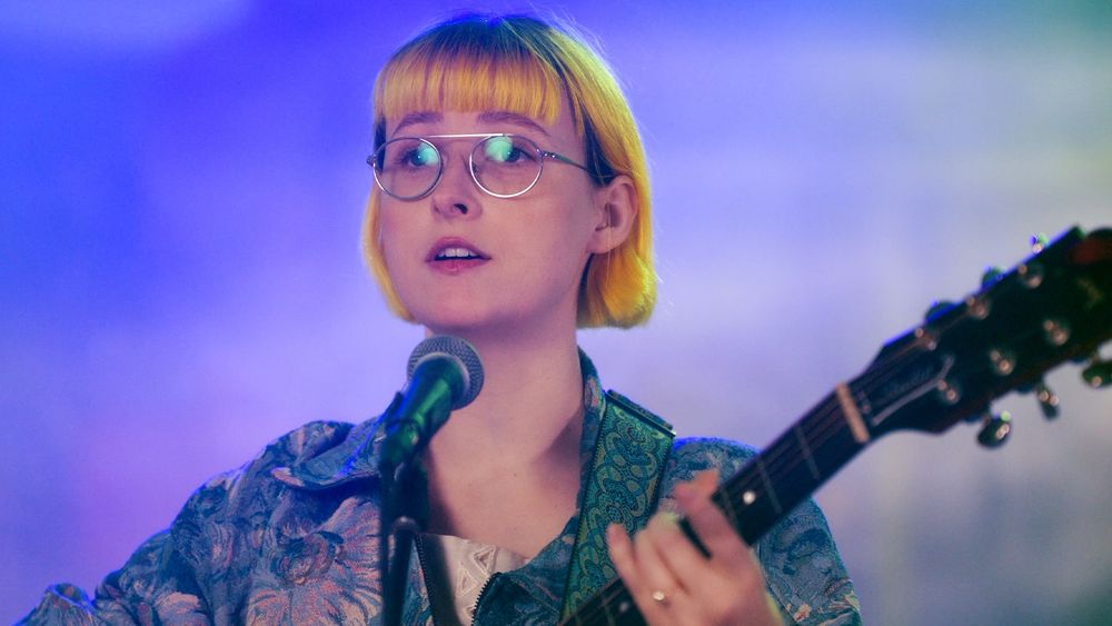 Tessa Violet in front of a microphone holding a guitar. 