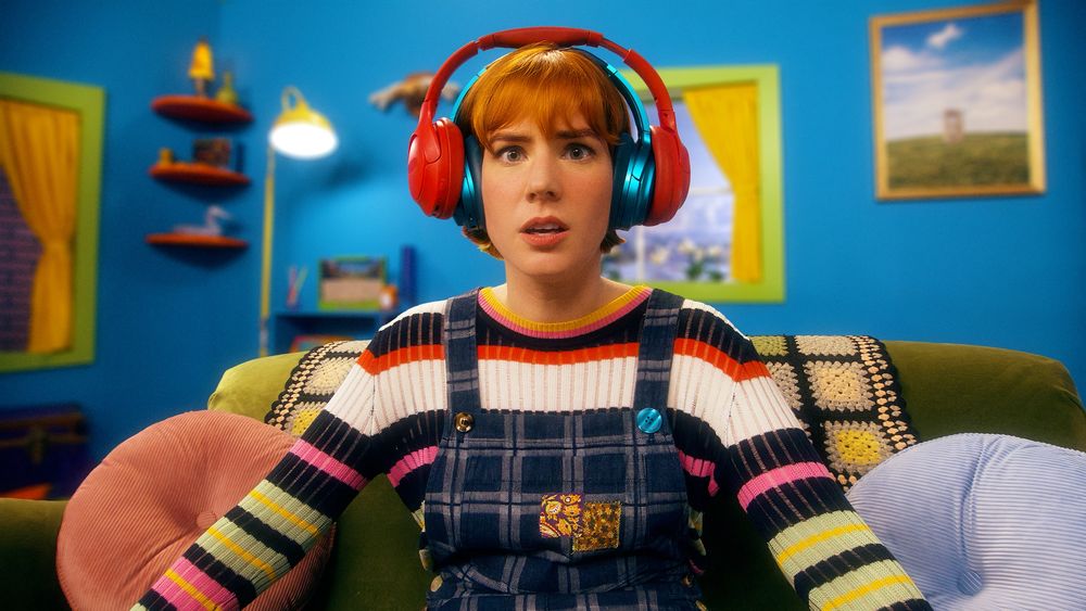Maggie Mae Fish wears two headphones, one blue and one red, and a striped sweater under overalls in her dream house. 