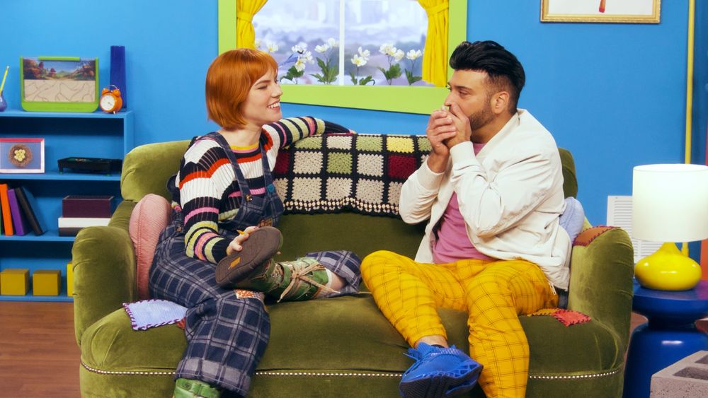 Maggie Mae Fish and Percy Rustomji sit on the green couch in Amy's living room. Percy is holding his hands in front of his mouth shocked. 
