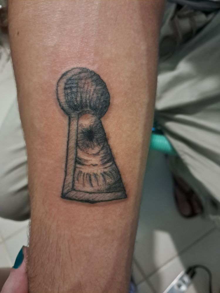 Tatuagem na parte interna do antebraço, tinha preta e traços finos, com o desenho de um olho espiando através de uma fechadura